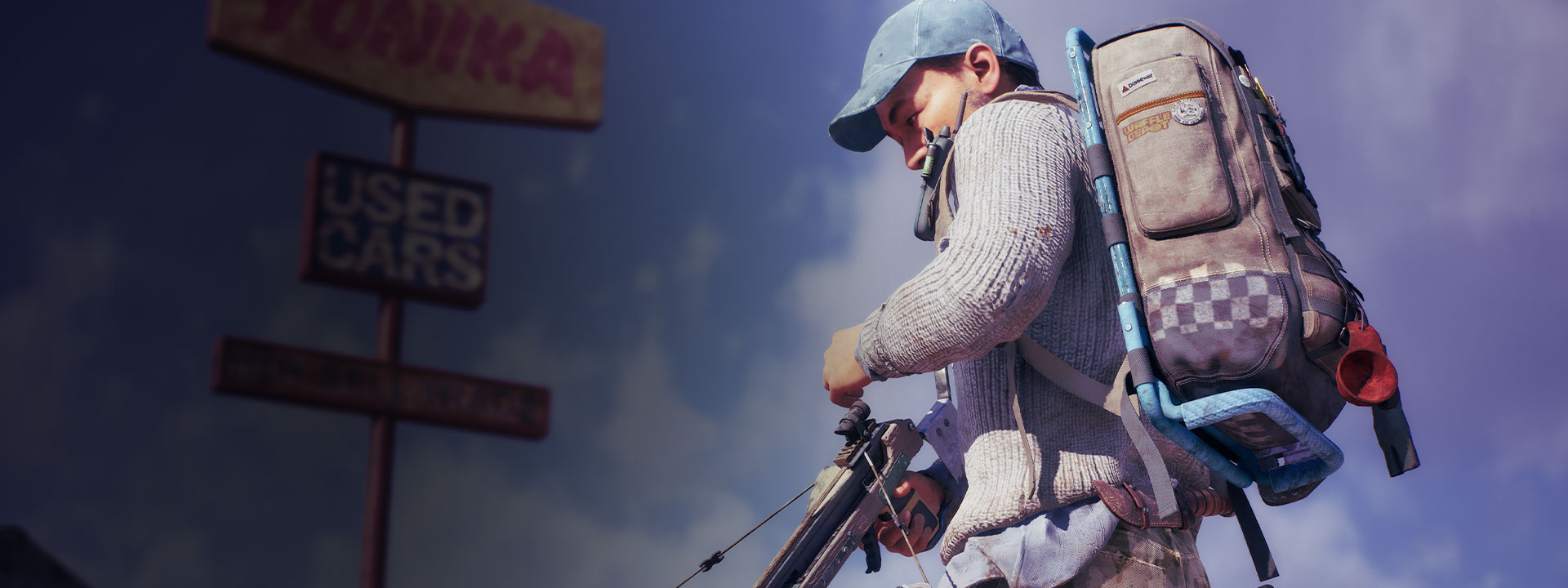 A man in a blue baseball cap loads up his crossbow under a large used car sign.