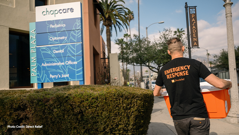 Photo Credit: Direct Relief, A person wearing an emergency response direct relief shirt walking on a Pasadena street with a box in their hands.