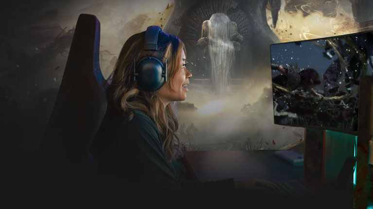 A woman sitting at her computer wearing a headset with Clair Obscur: Expeditions 33 gameplay on the monitor. 