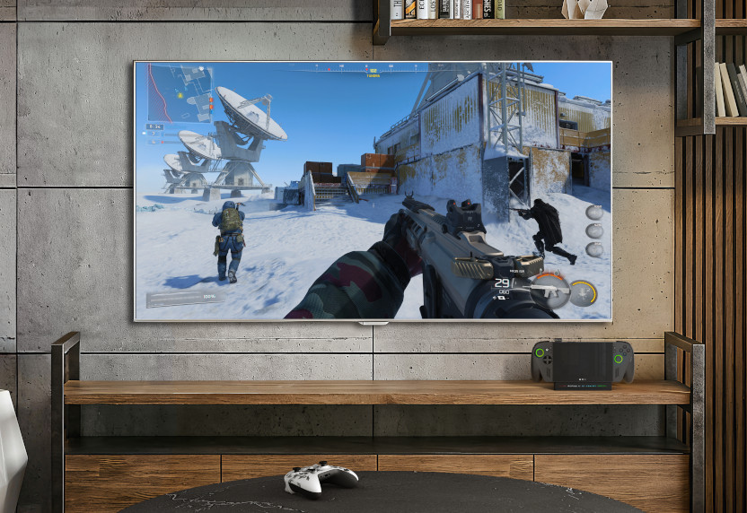 The ROG Xbox Ally X docked on a shelf below a TV with gameplay.