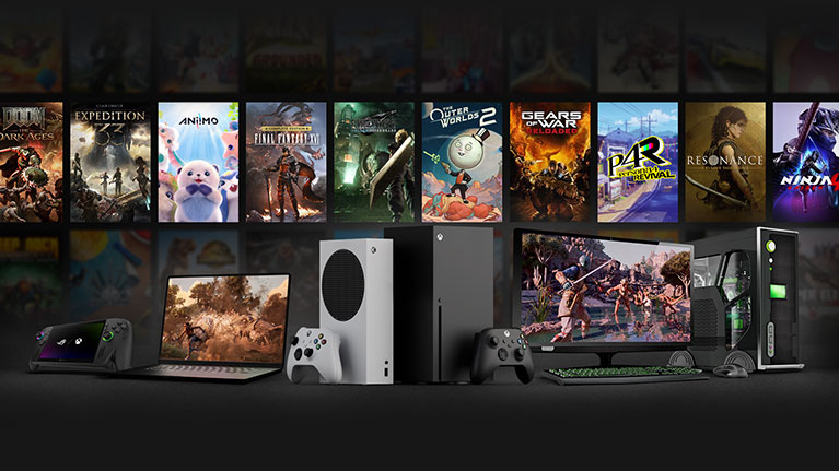 A handheld gaming device, laptop, Xbox Series S, Xbox Series X, Xbox wireless controllers, and a gaming PC with keyboard, mouse, monitor, and tower with an arrangement of game box art in the background.